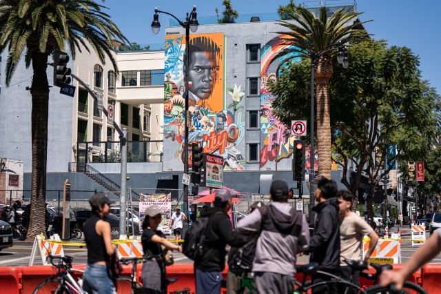 Bicyclists walking their bikes along a palm tree-lined street, in front of a building with a mural of Sidney Poitier on it.